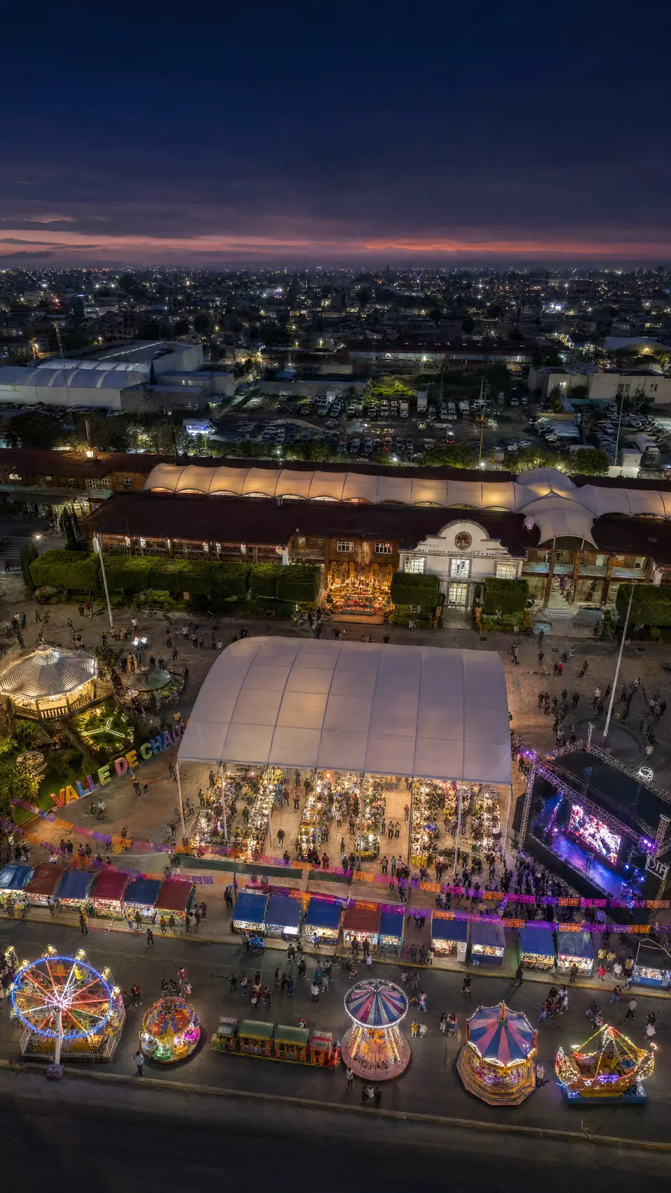 Ilustración aérea de la explanada al atardecer: al fondo el Palacio Municipal con sus arcos, al centro el domo blanco de productores, a la derecha el escenario, a la izquierda el kiosko y las letras «Valle de Chalco», y abajo la feria con la rueda de la fortuna y los juegos sobre la avenida.